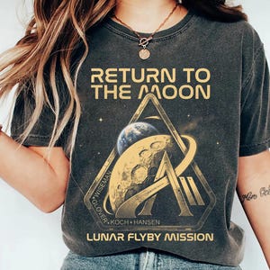 Op de afbeelding: Donkergrijs t-shirt met de tekst "RETURN TO THE MOON" boven een afbeelding van de maan, de aarde en een gestileerde "A". Het shirt heeft ook de tekst "LUNAR FLYBY MISSION" en de namen van de astronauten.