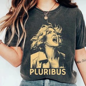 May include: Dark gray t-shirt featuring a yellow illustration of a woman with her mouth open and the word "PLURIBUS" in yellow block letters. The shirt is a crew neck and has short sleeves.