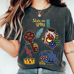 May include: Dark gray t-shirt with the text "Slay the Spire II" and various colorful graphic designs. The graphics include a key, a potion bottle with flames, a crown, a feather, and a blue creature.