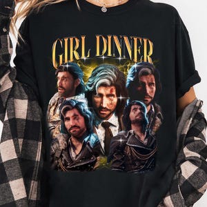 May include: Black t-shirt with the text "GIRL DINNER" in gold lettering. The shirt features a collage of images of a man with long hair. The shirt is worn by a person wearing a black and white plaid shirt.