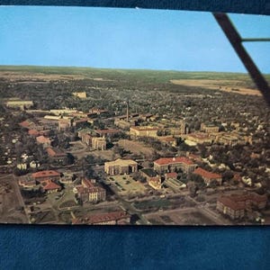 May include: Aerial view of a college campus with multiple buildings and red roofs. The image is a vintage postcard with a clear blue sky and a glimpse of an airplane wing in the upper right corner.