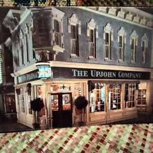 May include: A vintage postcard showing a black and white photograph of a building with the text "The Upjohn Company" above the shop front. The building has a sign that reads "Upjohn Pharmacy" and a large glass shop window displaying products.