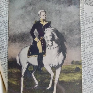 May include: A vintage postcard featuring a portrait of a man in a military uniform riding a white horse. The man is wearing a dark blue jacket with gold trim and a white sash. The horse is standing on a grassy field with trees in the background.