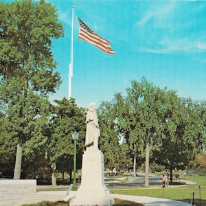 Puede incluir: Una estatua blanca de un hombre de pie sobre un pedestal en un parque. La estatua está frente a un asta de bandera con una bandera estadounidense ondeando en el viento. Hay árboles y hierba en el fondo.