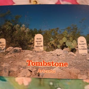 May include: A postcard featuring a scenic view of Tombstone, Arizona, with a clear blue sky. The image showcases several white tombstones with inscriptions, set against a backdrop of desert vegetation and rocky terrain. The word "Tombstone" is printed in red.