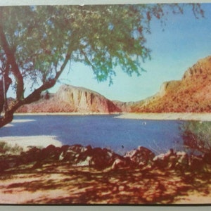 Könnte beinhalten: Eine Postkarte mit einer malerischen Landschaft, die einen See zeigt, der von Bergen umgeben ist, und einen großen Baum im Vordergrund. Der See ist leuchtend blau, und die Berge sind in einem warmen Braunton gehalten. Der Baum hat üppiges grünes Laub und Äste, die sich über den See erstrecken.