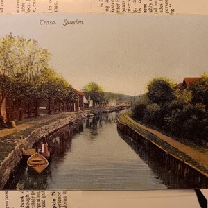 Puede incluir: Postal antigua que muestra una escena de canal en Trosa, Suecia. La imagen presenta una vía fluvial flanqueada por edificios y árboles, con un pequeño bote en el agua. La postal tiene un tono sepia, evocando nostalgia.