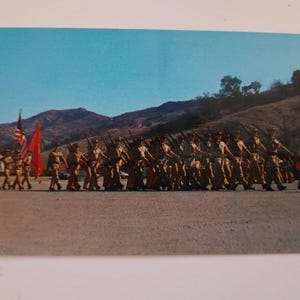 May include: Vintage postcard featuring a military parade. Soldiers in uniform march in formation, carrying rifles. The American flag and a red flag are held by soldiers at the front. Mountains and a blue sky form the backdrop.