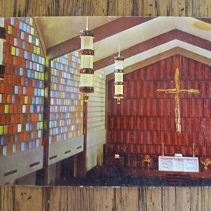 Könnte beinhalten: Eine Vintage-Postkarte, die das Innere einer Kirche mit Buntglasfenstern, einem großen Holzkreuz und einem weißen Altar zeigt. Die Kirche hat ein modernes Design mit einer hohen Decke und einem großen, offenen Raum.