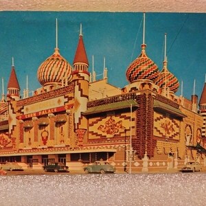 Puede incluir: Una postal vintage que representa el Corn Palace en Mitchell, Dakota del Sur. El edificio está decorado con murales coloridos y diseños intrincados, con una mezcla de patrones geométricos y figuras estilizadas. El edificio tiene un estilo arquitectónico único con múltiples torres y cúpulas.