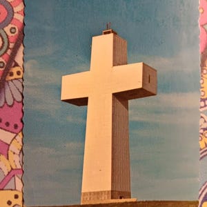 Puede incluir: Una postal vintage con una gran cruz blanca sobre un cielo azul. La cruz es alta y prominente, situada sobre una colina cubierta de hierba. La postal tiene un borde festoneado y un borde floral colorido.