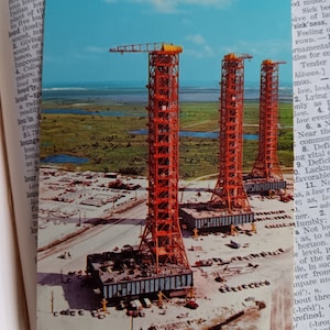 May include: Three large red metal structures stand on a flat, sandy surface. The structures are tall and narrow, with multiple levels and beams. The structures are set against a blue sky with white clouds.