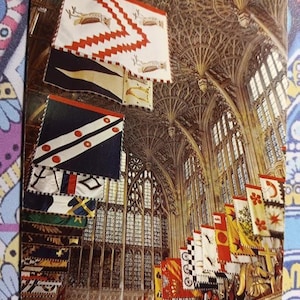 Puede incluir: Una fotografía del interior de una catedral con numerosas banderas coloridas colgadas del techo. Las banderas muestran varios diseños heráldicos, incluyendo zigzags, ciervos y patrones geométricos. La arquitectura presenta altas ventanas arqueadas y detalles ornamentales en el techo.