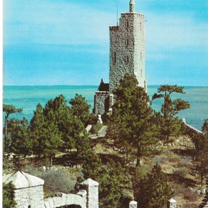May include: A stone tower with a pointed roof stands on a hill overlooking a body of water. The tower is surrounded by trees and a stone gate with a black metal fence.