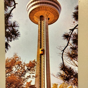 Peut inclure: Carte postale vintage représentant la tour Skylon du Centre International de Niagara. La tour a un sommet distinctif en forme de champignon avec un motif à carreaux. Un ascenseur jaune monte la tour. L'image est encadrée par des branches d'arbres et du feuillage.