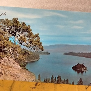 May include: A postcard featuring a scenic view of a lake with a small island in the middle. The lake is surrounded by trees and mountains. The sky is blue with white clouds.