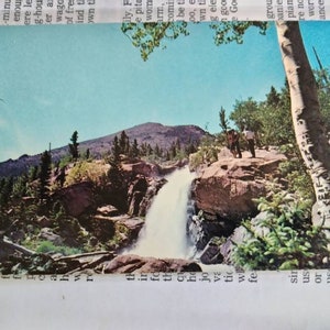 Puede incluir: Una postal con una cascada pintoresca que cae por un acantilado rocoso en un bosque verde exuberante. La cascada está rodeada de árboles y rocas, y el cielo es de un azul brillante. Dos personas están de pie en una cornisa rocosa con vistas a la cascada.