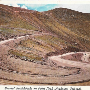 May include: A winding dirt road with several switchbacks on Pikes Peak Highway in Colorado. The road is surrounded by brown hills and a blue sky with white clouds.
