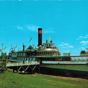 May include: A vintage white and black steamboat moored at a pier. The boat has a red and black funnel and a white railing. The boat is surrounded by trees and a blue sky.