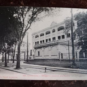 May include: Black and white vintage postcard featuring a large building with multiple windows and a decorative facade. Trees line the street in front of the building. The text on the postcard reads "243 Cochinchine - Saigon - Caserne des Marins."