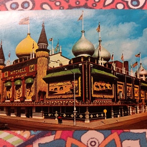 Könnte beinhalten: Eine Vintage-Postkarte, die das Mitchell Corn Palace in Mitchell, South Dakota, zeigt. Das Gebäude ist mit bunten Wandgemälden verziert und hat ein fantasievolles, märchenhaftes Aussehen. Der Text "Mitchell Corn Palace" und "64" ist auf dem Gebäude sichtbar.