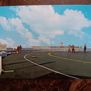 May include: A vintage postcard showing a black and white photograph of a large aircraft carrier docked at a pier. The postcard has a blue sky with white clouds and the text "Chesapeake Bay Bridge Tunnel" on a white sign in the foreground.