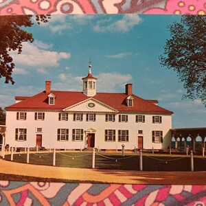May include: Vintage postcard featuring a white colonial-style building with a red roof and multiple windows. The building has a central cupola and is surrounded by trees and a blue sky with clouds. A fence and a pathway are in the foreground.