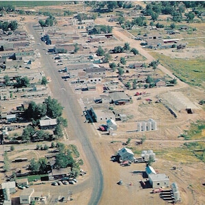 May include: An aerial view of a small town with a long, straight road running through the center. The town is surrounded by fields and trees. There are several buildings on either side of the road, including houses, businesses, and a church.