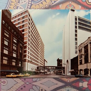 May include: A vintage postcard featuring a view of a city street with tall buildings. The buildings are a mix of brick and concrete, and they are all connected by a pedestrian bridge. The sky is blue and there are clouds in the distance.