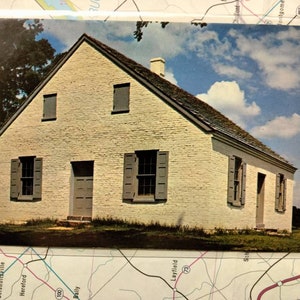 May include: A white brick building with a gabled roof and six windows. The building has a wooden door and is surrounded by green grass.