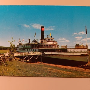May include: A vintage postcard depicting a white and black steamboat moored at a wooden jetty. The boat has a red and black funnel and several flags flying from its masts. There are people on the deck and the jetty. The postcard is a vintage image with a faded appearance.