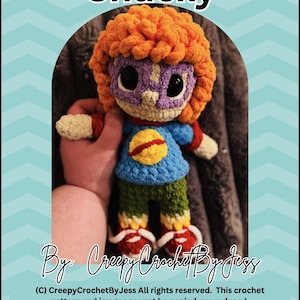 Könnte beinhalten: Eine handgefertigte Häkelpuppe namens Chucky mit leuchtend orangefarbenem lockigem Haar, einer violetten Maske, einem blauen Oberteil mit gelbem kreisförmigem Emblem, grüner Hose und roten Schuhen. Der Text Chucky, By: CreepyCrochetByJess ist zu sehen.