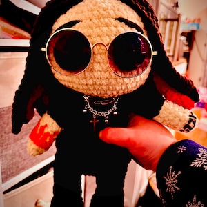 May include: A handmade crochet doll with a cream-colored face, black hair, and black clothing. The doll wears round gold-rimmed sunglasses and a silver chain necklace with a cross pendant. The doll's arm has a red and cream-colored design.