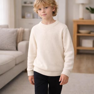May include: A cream-colored sweater with a crew neck and ribbed cuffs and hem. The sweater is worn with dark blue pants. The image is taken indoors, with a couch and wooden furniture in the background.
