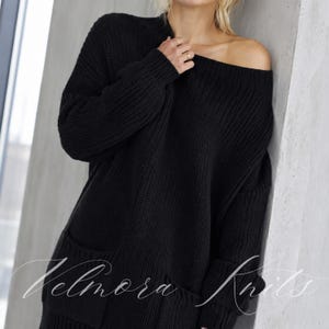 May include: A black long-sleeved sweater featuring a chunky knit texture, a wide boat neckline, and two front pockets. The garment displays the script text Velmora Knits overlaid across the lower portion of the fabric in a white cursive font.