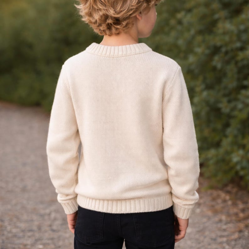 May include: Cream-colored knit sweater with a crew neck and long sleeves. The sweater is made of a soft, textured material. The person is wearing black pants. The background is a blurred green and gray.