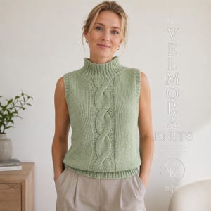 May include: A sage green sleeveless knit sweater vest featuring a mock neck and a prominent vertical cable knit pattern down the center. The text VELMORA KNITS is printed on the right side in a decorative white font with a small logo below.