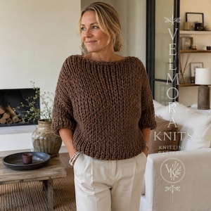 Pode incluir: Uma camisola de malha grossa em cor castanho escuro sólido com um decote barco largo e mangas três quartos. A peça tem um padrão de malha espesso e texturizado com um ajuste relaxado. O texto VELMORA KNITS KNITTING PATTERNS é visível.