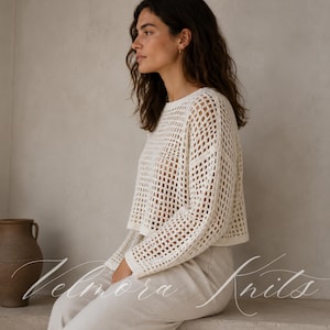 May include: A cream-coloured, open-weave knit sweater with a crew neck and long sleeves. The sweater is worn with light-coloured trousers. The image also includes the text "Velmora Knits" in a cursive font.