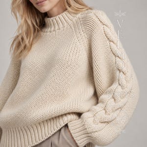 Op de afbeelding: Een crèmekleurige trui met lange mouwen, een opstaande kraag en een grove breistructuur. De mouwen hebben een opvallend verticaal kabelbreipatroon aan de buitenkant. De tekst VELMORA KNITS is zichtbaar aan de rechterkant van de afbeelding.