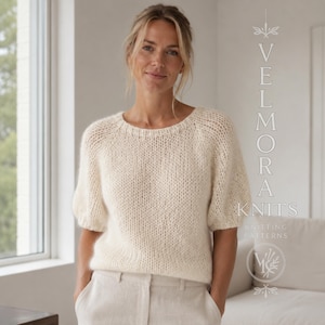 Può includere: Un maglione in maglia color crema a maniche corte con scollo rotondo e una lavorazione a punto largo e testurizzato. Il testo VELMORA KNITS KNITTING PATTERNS è visibile sul lato destro, insieme a un logo circolare con un disegno vegetale stilizzato.