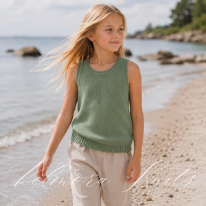 Könnte beinhalten: Ein Kind trägt ein ärmelloses grünes Strickoberteil und eine beige Hose an einem Sandstrand. Das Oberteil hat einen gerippten Ausschnitt und Saum. Das Meer und die Küste sind im Hintergrund zu sehen. Der Text "Velmora Knits" ist kursiv am unteren Bildrand geschrieben.