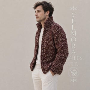 Può includere: Un cardigan a maglia grossa in un misto variegato di filato bordeaux scuro e crema, caratterizzato da un collo alto e maniche lunghe. Il capo è indossato sopra una camicia color crema tinta unita e pantaloni chiari. Il testo a destra recita VELMORA KNITS.