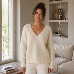 Könnte beinhalten: Ein cremefarbener Langarmpullover mit V-Ausschnitt sowie gerippten Bündchen und Saum. Das Strickkleidungsstück hat eine weiche, flauschige Textur und wird mit einer hellen Hose getragen. Der Text VELMORA KNITS KNITTING PATTERNS ist sichtbar.