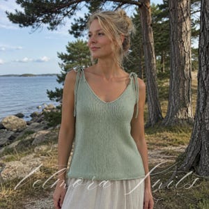 Puede incluir: Un top de punto verde claro con cuello en V y tirantes anudados. El top se lleva con una falda de color claro. El texto "Elmora Knits" es visible en la parte inferior de la imagen. El fondo incluye árboles, agua y un cielo azul.