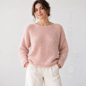 May include: A soft, pale pink long-sleeved sweater with a wide boat neckline and a loose, ribbed knit texture. The garment features dropped shoulders and a relaxed fit, paired with plain white trousers. Text on the right reads VELMORA KNITS.