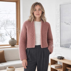 May include: A dusty rose, open-front knit cardigan with long sleeves and a textured stitch pattern. The garment is worn over a plain white shirt and dark gray trousers. The text VELMORA KNITS KNITTING PATTERNS is visible on the right side.