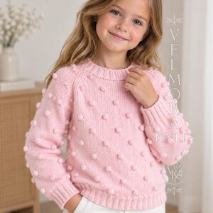 May include: A light pink long-sleeve knit sweater featuring a textured bobble pattern across the front and sleeves, with a ribbed crew neckline, cuffs, and hem. The text VELMORA KNITS is printed vertically on the right side of the image.