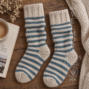 Könnte beinhalten: Ein Paar gestrickte Socken mit einem Streifenmuster in Beige und Dunkelblau. Die Socken haben ein geripptes Bündchen am oberen Rand sowie eine verstärkte Ferse und Spitze. Der Text VELMORA KNITS ist rechts im Bild zu sehen.