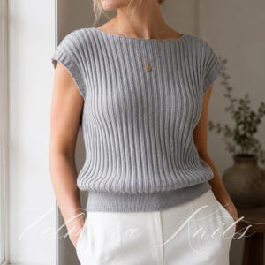 May include: A light gray sleeveless top featuring a vertical ribbed knit texture, a wide boat neckline, and a fitted hemline. The garment is worn with white trousers and a delicate gold necklace with a small circular pendant resting on the chest.